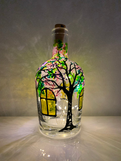 The secret garden is seen through windows that are aglow with yellow panes.  Trees with colourful leaves surround. Painted on a recycled bottle with acrylic paint. The bottle is lit with fairy lights from within.